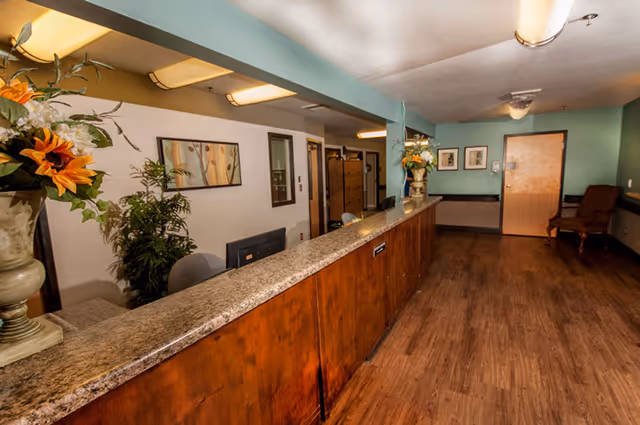 Reception area with a long granite countertop desk, wooden paneling, and two large flower arrangements in vases. Behind the desk are office chairs, a computer monitor, framed artwork on the wall, and some plants. The floor is wood, and there is a door and a chair against a teal-colored wall in the background.