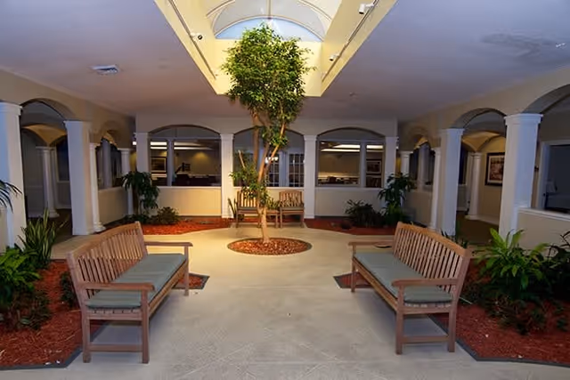 Indoor courtyard area with a skylight ceiling, featuring a central tree surrounded by mulch and plants. There are wooden benches with cushions arranged around the tree, and white columns and arches frame the space with windows and doorways in the background.