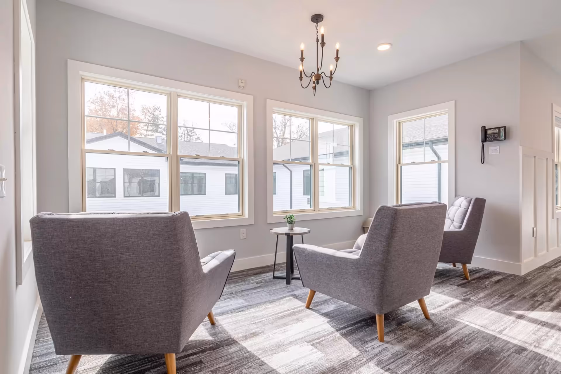 A bright sitting area with three gray upholstered armchairs arranged around a small round table with a small plant on it. The room has large windows letting in natural light, light gray walls, a modern chandelier, and a patterned carpeted floor.