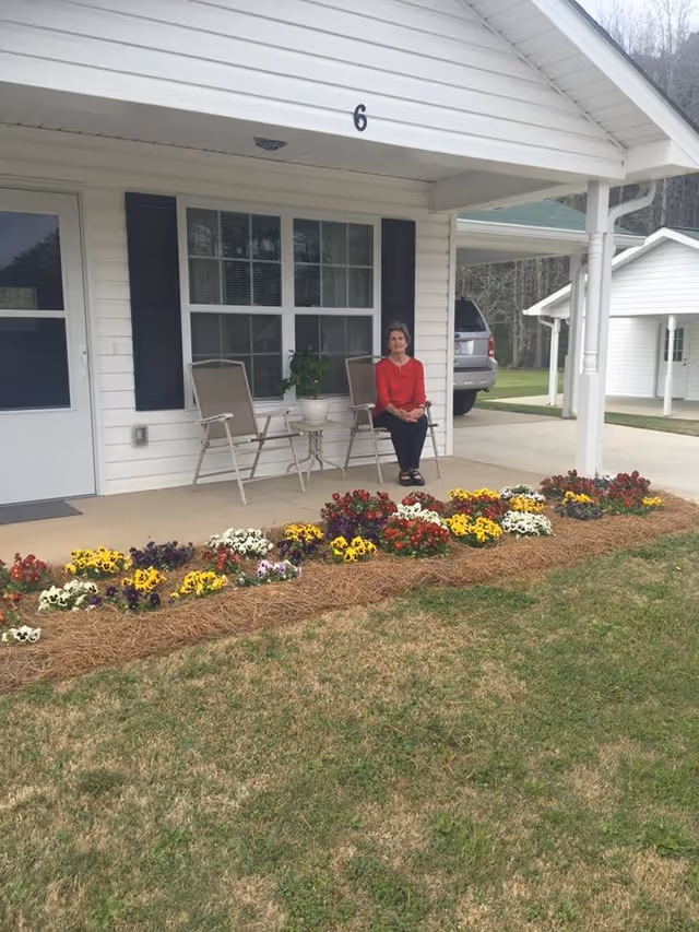 A woman in a red top sitting on a chair on the porch of a white house with black shutters. There are two chairs and a small table with a potted plant on the porch. In front of the porch is a flower bed with colorful flowers and pine straw mulch. A silver vehicle is parked in the driveway next to the house.