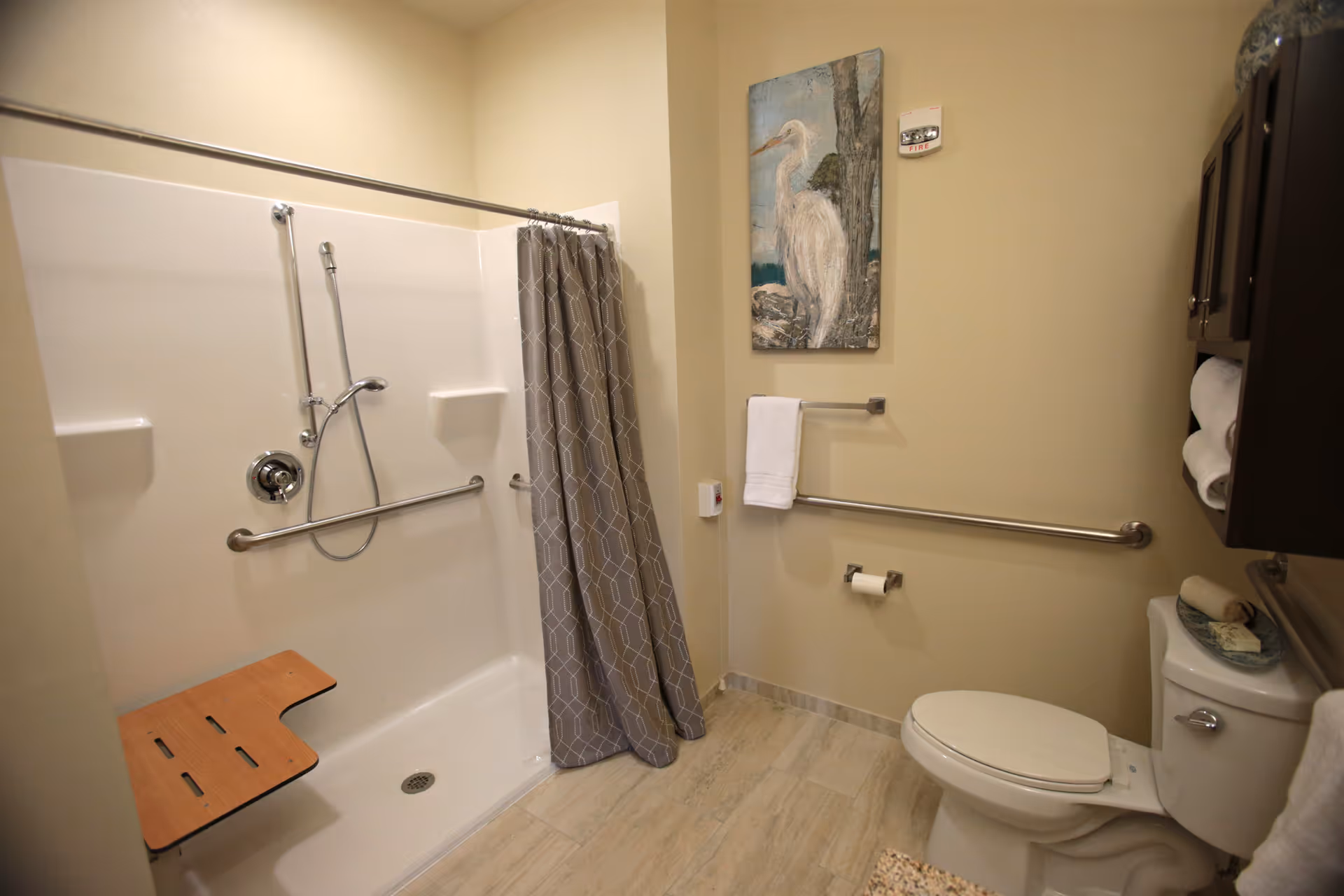 A bathroom with a walk-in shower featuring a handheld showerhead and a wooden fold-down seat. There is a gray patterned shower curtain, a white toilet with a roll of toilet paper on the wall, and a towel hanging on a bar. Above the towel bar is a painting of a white bird standing near a tree. The floor is tiled, and there is a dark wooden cabinet with rolled white towels on the right wall.