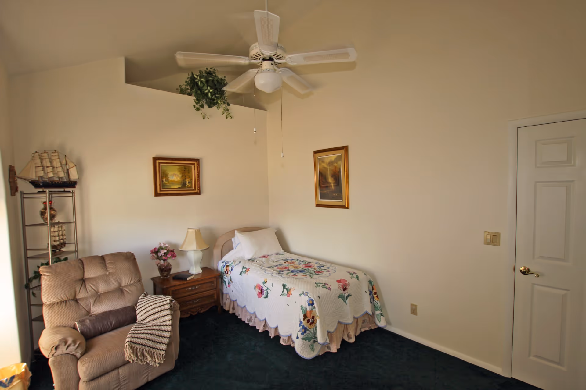 A small bedroom with a single bed covered in a floral quilt, a wooden nightstand with a lamp and a vase of flowers, a beige recliner chair with a striped blanket draped over it, a metal shelf with decorative items including a model ship, two framed paintings on the walls, a ceiling fan, and a closed white door.