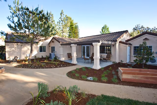 Exterior view of a single-story building with a tiled roof and a covered patio area furnished with chairs and a table. The surrounding area features landscaped garden beds with small plants and trees under a clear blue sky.