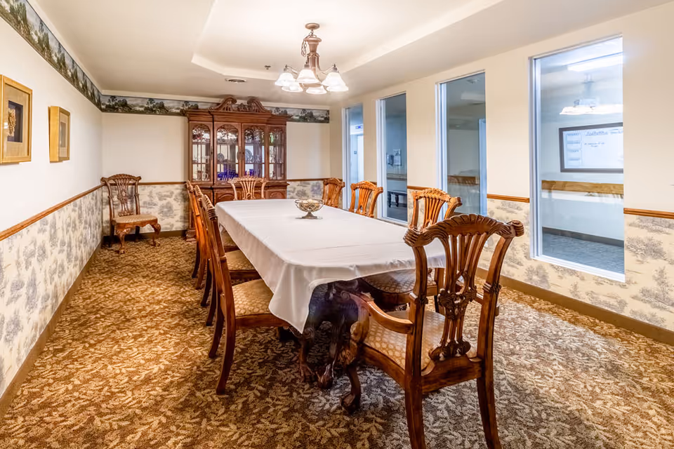A formal dining room with a long rectangular table covered with a white tablecloth surrounded by wooden chairs with cushioned seats. The room has patterned carpet and wallpaper with a nature motif, a wooden china cabinet at the far end, and three large windows on one side showing an adjacent hallway.