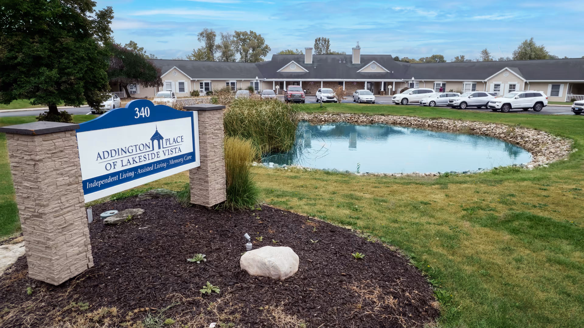 Exterior view of Addington Place of Lakeside Vista facility with a pond in the foreground, a landscaped area with a sign displaying the facility name and services, and a row of parked cars in front of the building under a partly cloudy sky.