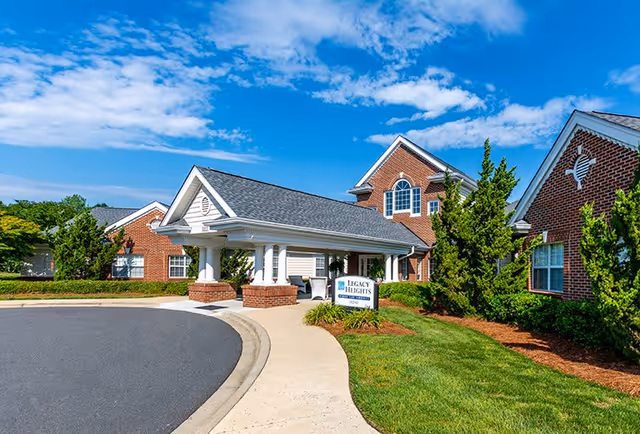 Exterior view of Legacy Heights Senior Living Community featuring a brick building with white columns at the entrance, a curved driveway, green shrubs, and a clear blue sky with some clouds.