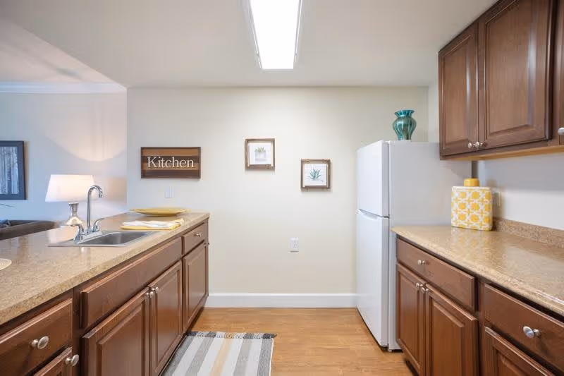 A clean kitchen with wooden cabinets and beige countertops. There is a white refrigerator on the right side and a sink with a faucet on the left countertop. On the wall, there are three framed decorations, one of which says 'Kitchen'. The floor is wooden with a striped rug in front of the sink. A lamp and part of a living room are visible in the background.