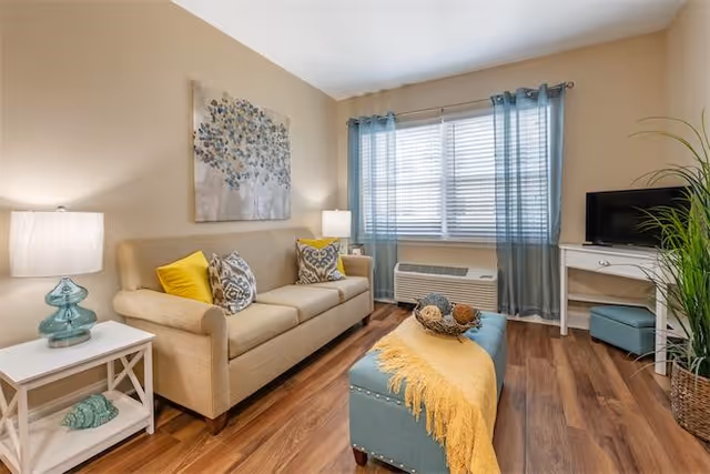 Cozy living room with a beige sofa, patterned pillows, a blue ottoman with a yellow throw, a TV on a white desk, and a window with blue curtains.