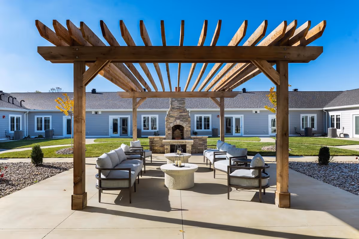 Outdoor seating area at Cedarhurst Senior Living of Nicholasville featuring cushioned chairs and sofas arranged around a stone fireplace under a wooden pergola, with a clear blue sky and single-story building in the background.