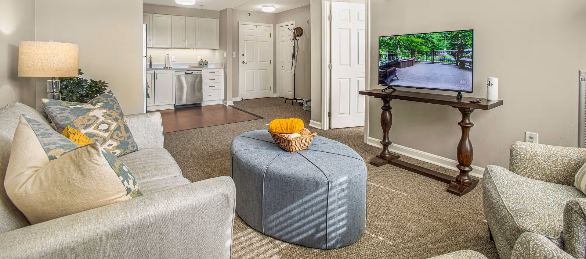 Well-lit living room with a sofa, patterned armchair, round ottoman, TV on a console and a small kitchenette visible in the background.