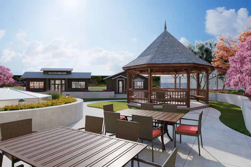 Outdoor seating area with multiple tables and chairs with red cushions, a wooden gazebo with a pointed roof, surrounded by green grass, flowering trees, and a clear blue sky.