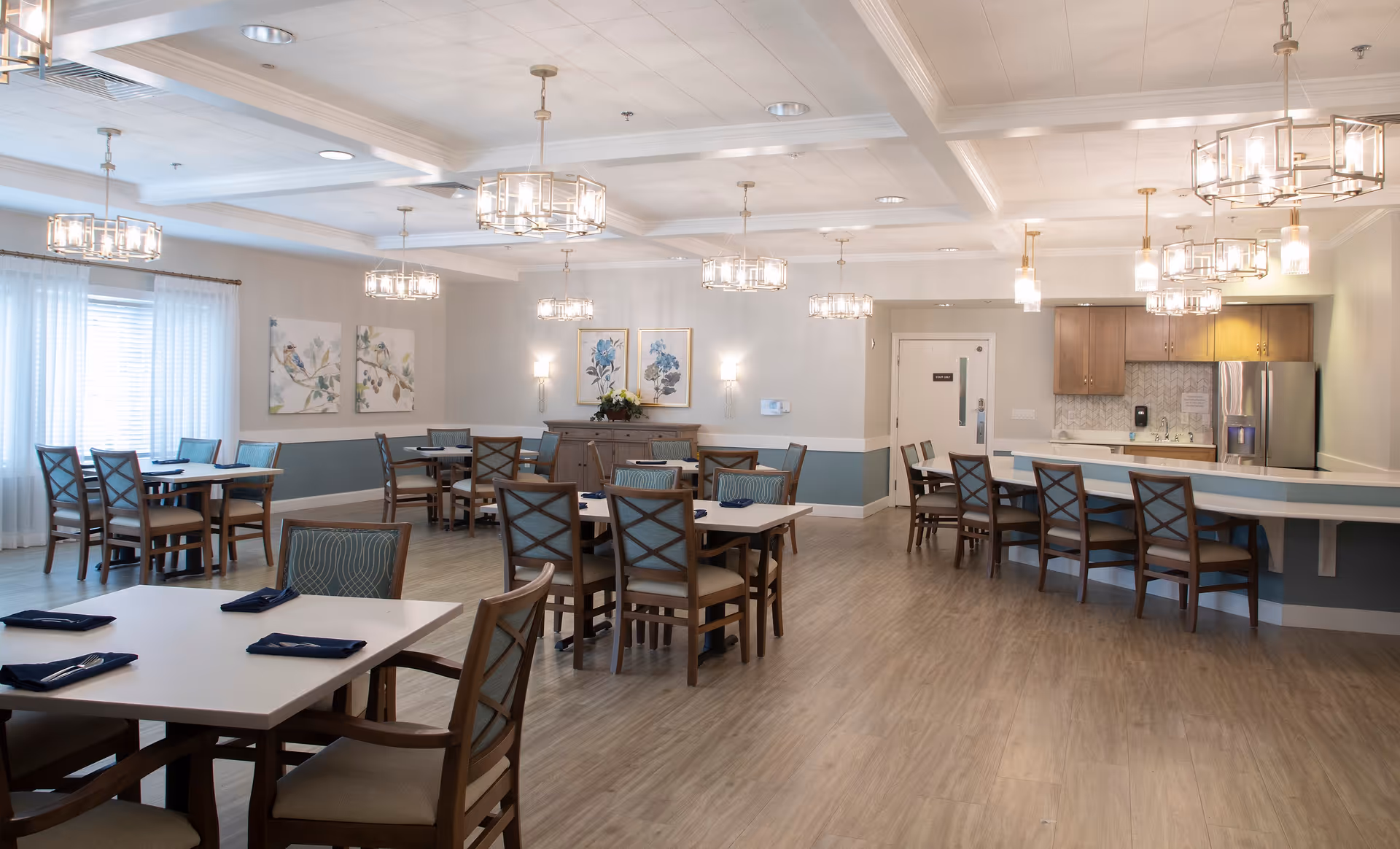 A spacious, well-lit dining room with multiple tables and chairs arranged neatly. The room features modern pendant lights hanging from the ceiling, light-colored walls with blue accents, framed floral and bird artwork, and a kitchen area with a counter and bar stools. The floor is wood-style, and windows with sheer white curtains allow natural light to enter.
