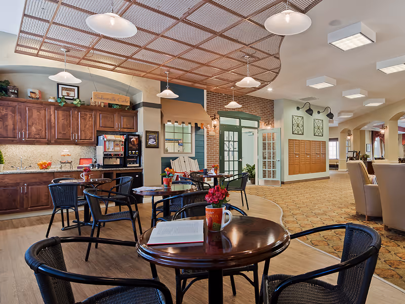 Bright communal cafe-style dining area with round tables and chairs, a kitchenette and decorative ceiling details.