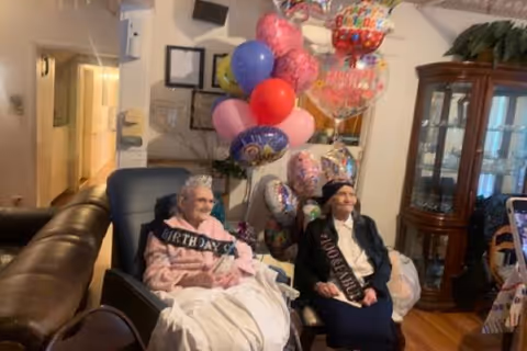 Two elderly women sitting in chairs indoors, both wearing birthday sashes and tiaras. Behind them is a bunch of colorful balloons, including some with birthday messages. The room has wooden flooring, a glass display cabinet, and some furniture.
