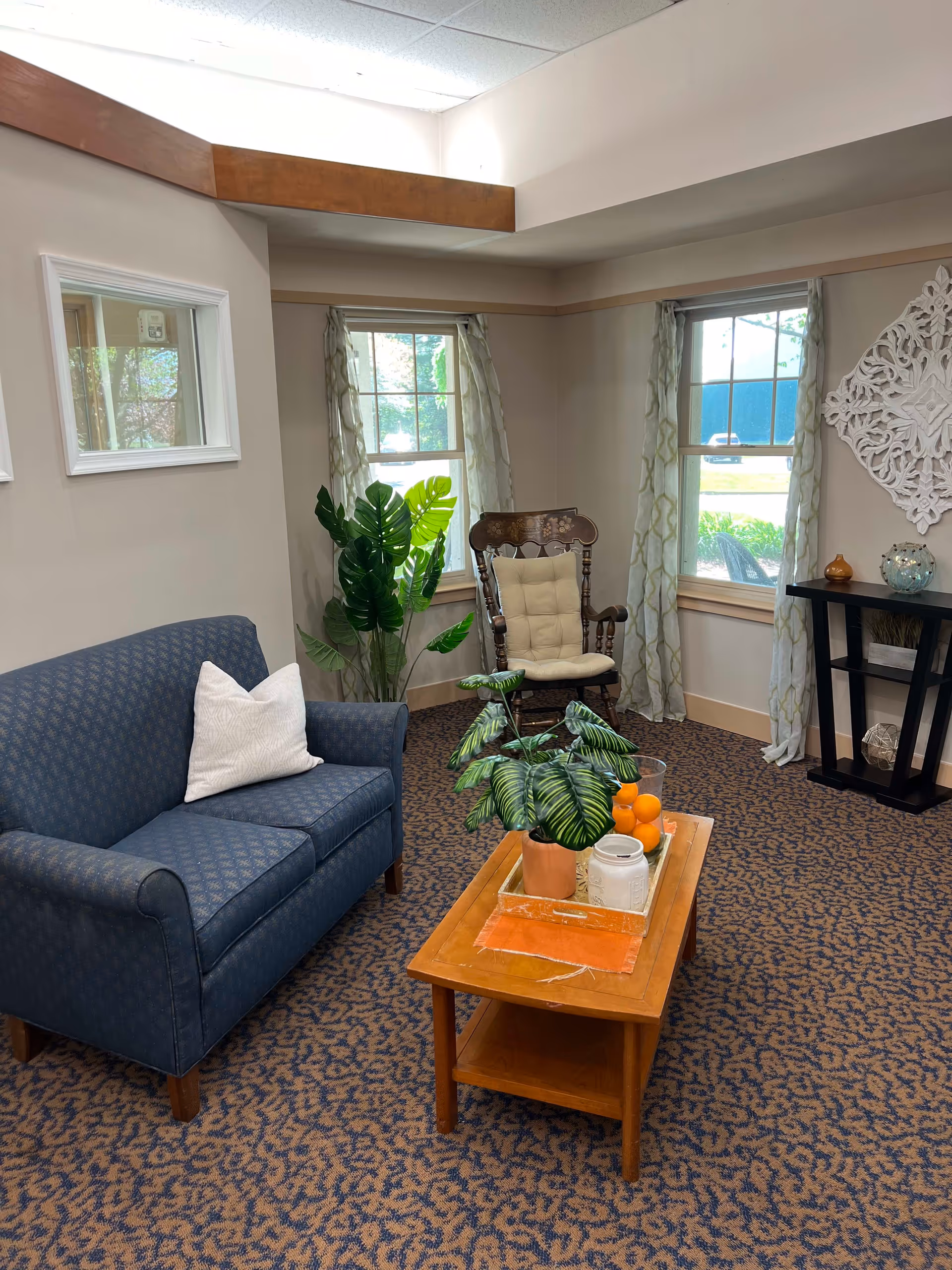 A cozy living room area with a blue patterned loveseat with a white pillow, a wooden rocking chair with a beige cushion, and a wooden coffee table with a potted plant, a tray, and a bowl of oranges. The room has two windows with light curtains, a decorative white wall piece, a small black console table with decorative items, and a patterned carpet.