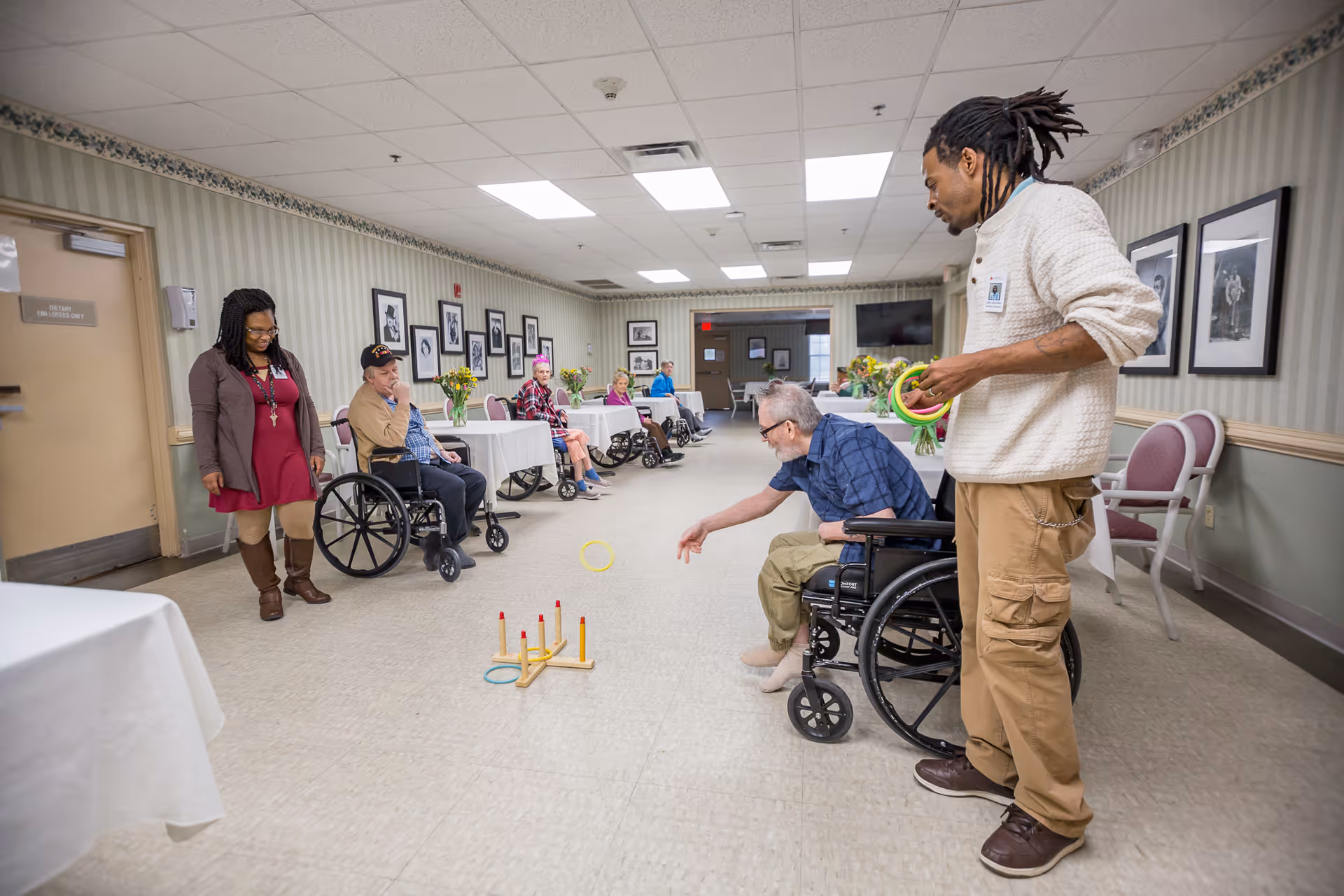 A group of elderly residents and staff in a long room at Maplewood Health Care Center. One elderly man in a wheelchair is playing a ring toss game with the assistance of a staff member holding rings. Other residents, some in wheelchairs, and staff watch and participate in the activity. The room has tables with white tablecloths and flowers, framed pictures on the walls, and a tiled floor.