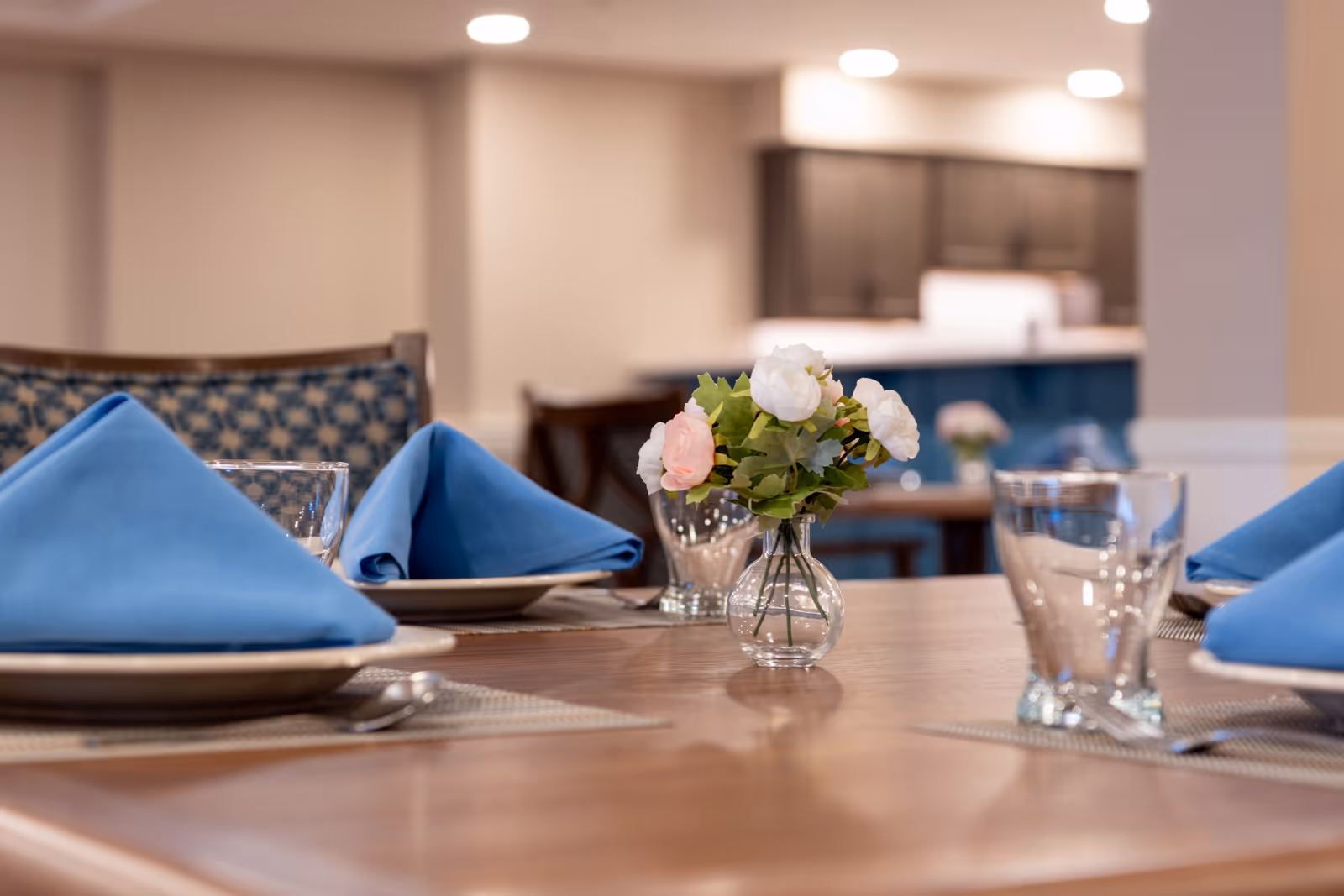 Close-up view of a dining table set with blue folded napkins on plates, clear drinking glasses, and a small vase with white and pink flowers in the center. The background shows a softly lit interior with chairs and kitchen cabinets.