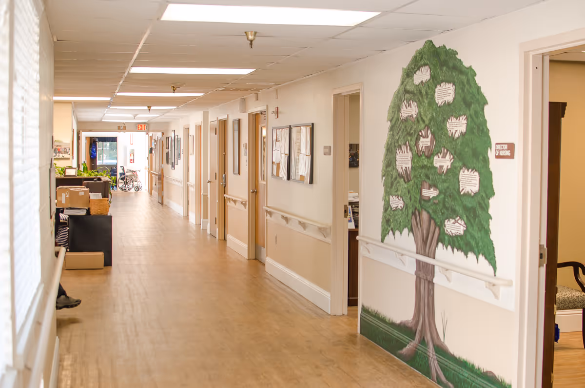 A long, well-lit hallway in a senior living facility with wooden floors and handrails along the walls. Several doors line the hallway, and a large mural of a tree with labeled leaves is painted on the right wall. There is a sign next to the mural that reads 'Director of Nursing.' At the far end of the hallway, a wheelchair is visible near an exit door.