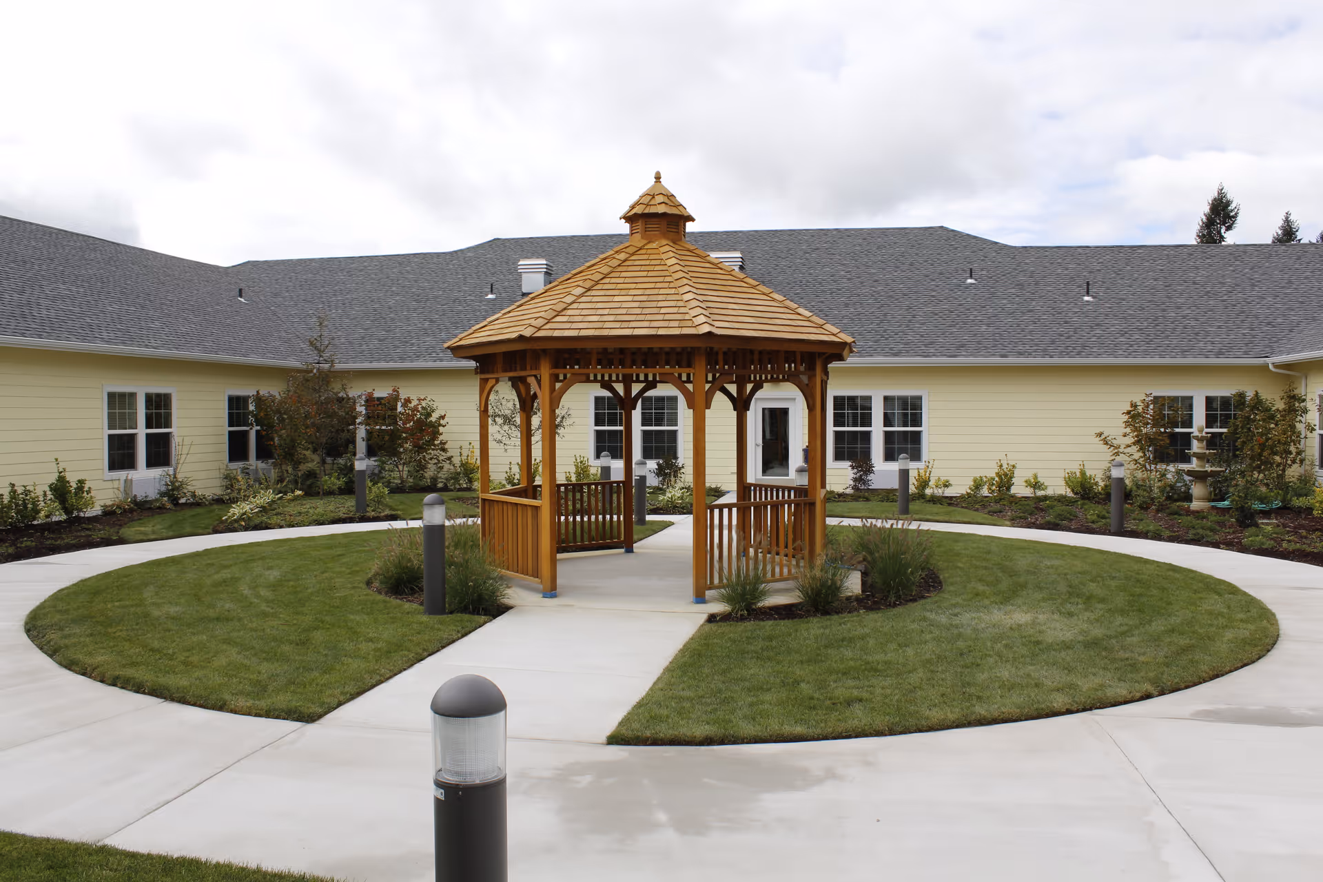 A wooden gazebo with a shingled roof is situated in the center of a circular concrete pathway surrounded by well-maintained green grass and landscaped plants. The background features a single-story yellow building with multiple white-framed windows and a gray shingled roof under a cloudy sky.