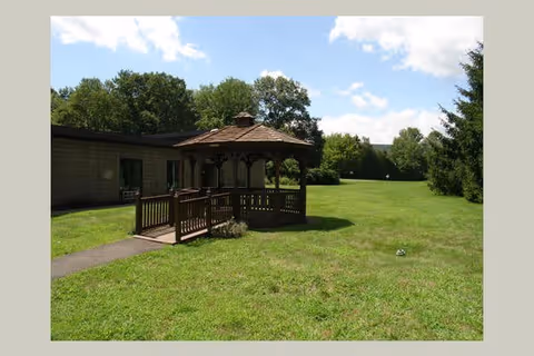 A wooden gazebo with a shingled roof situated on a grassy lawn next to a building. Trees and a partly cloudy sky are visible in the background.