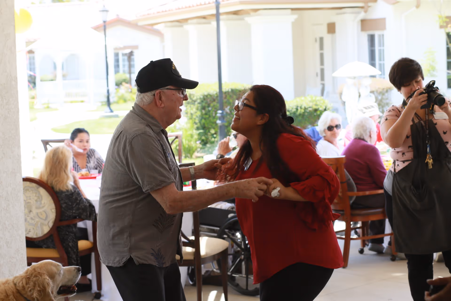 An elderly man and a woman in a red blouse are holding hands and smiling at each other outdoors at a senior living community. In the background, several elderly people are seated at tables, and a woman is taking photos with a camera. A golden retriever dog is also visible near the elderly man.
