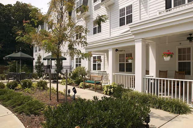 Outdoor patio area of a senior living facility with white siding buildings, benches, hanging flower pots, green umbrellas over tables, and landscaped garden beds with shrubs and small trees.
