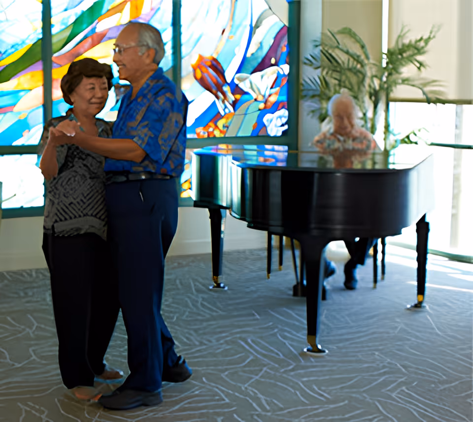 An elderly couple dancing together in a room with a grand piano and a colorful stained glass window in the background. Another elderly person is seated at the piano, playing music. The room has large windows and plants, creating a bright and welcoming atmosphere.
