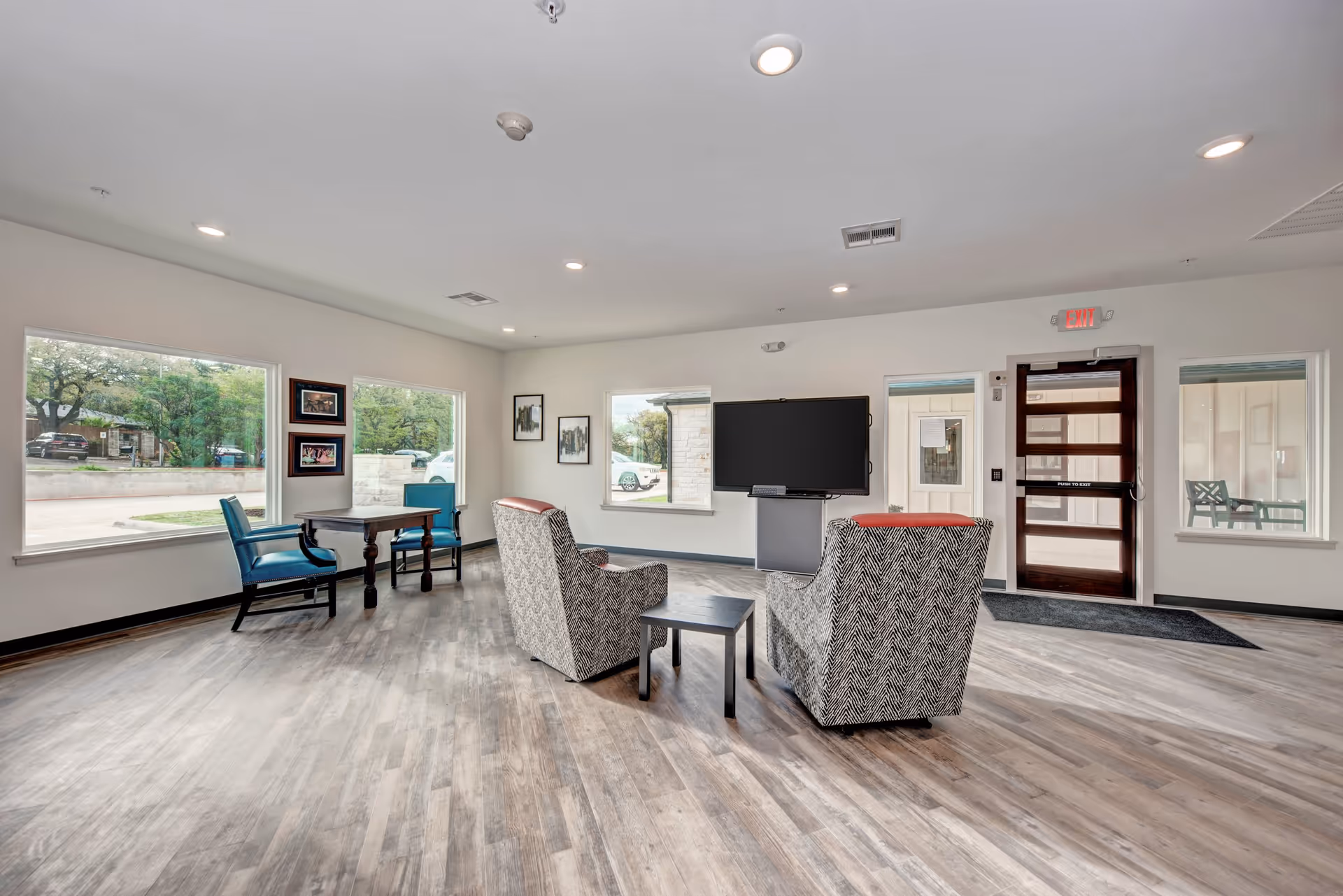A spacious and bright common area with large windows showing outdoor greenery. The room features two patterned armchairs with red headrests facing a large flat-screen TV mounted on a stand. There is a small black side table between the chairs. In the corner, there is a wooden table with two blue upholstered chairs. The floor is wood-style laminate, and the walls are painted white. An exit door with glass panels is visible on the right side of the room.