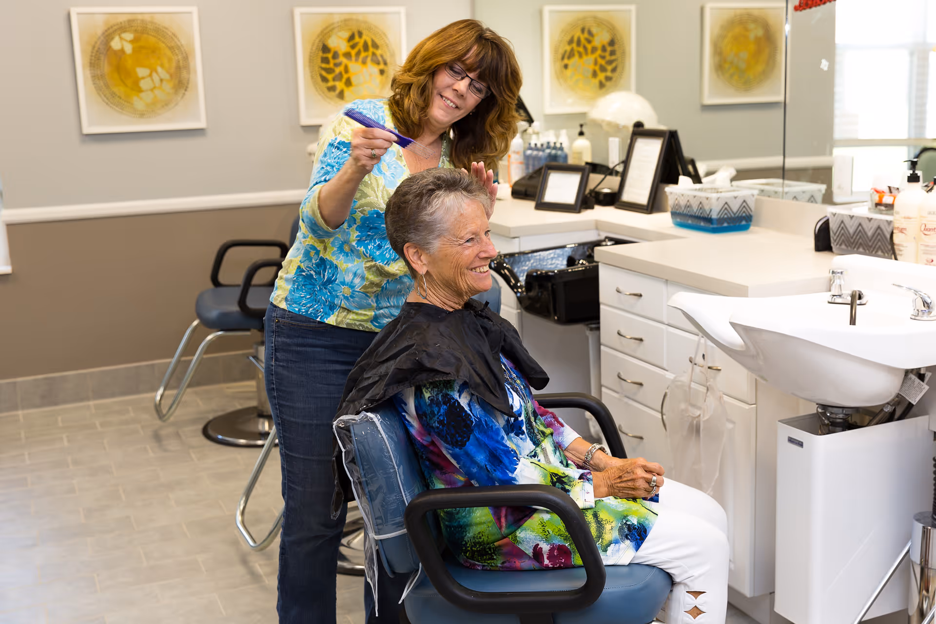 A senior woman sitting in a salon chair smiling while a hairstylist cuts her hair in a well-lit salon area with mirrors, a sink, and hair care products visible.