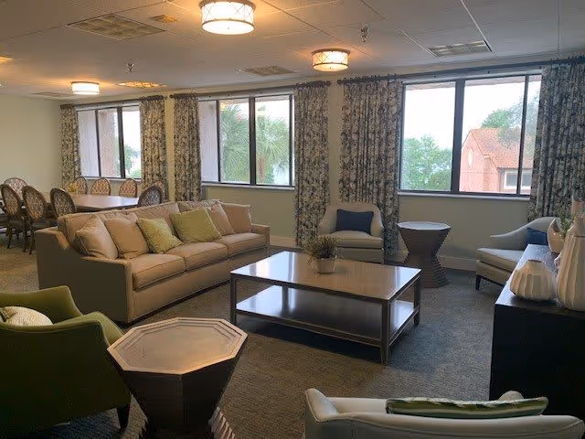 A spacious living room area with a beige sofa, several armchairs, a wooden coffee table with a small plant, and a dining table with chairs near large windows with patterned curtains. The room is well-lit with ceiling lights and has a carpeted floor.