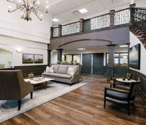 Two-story elegant lobby with sofas, armchairs, a chandelier, hardwood floors, and an upper balcony with ornate railing.