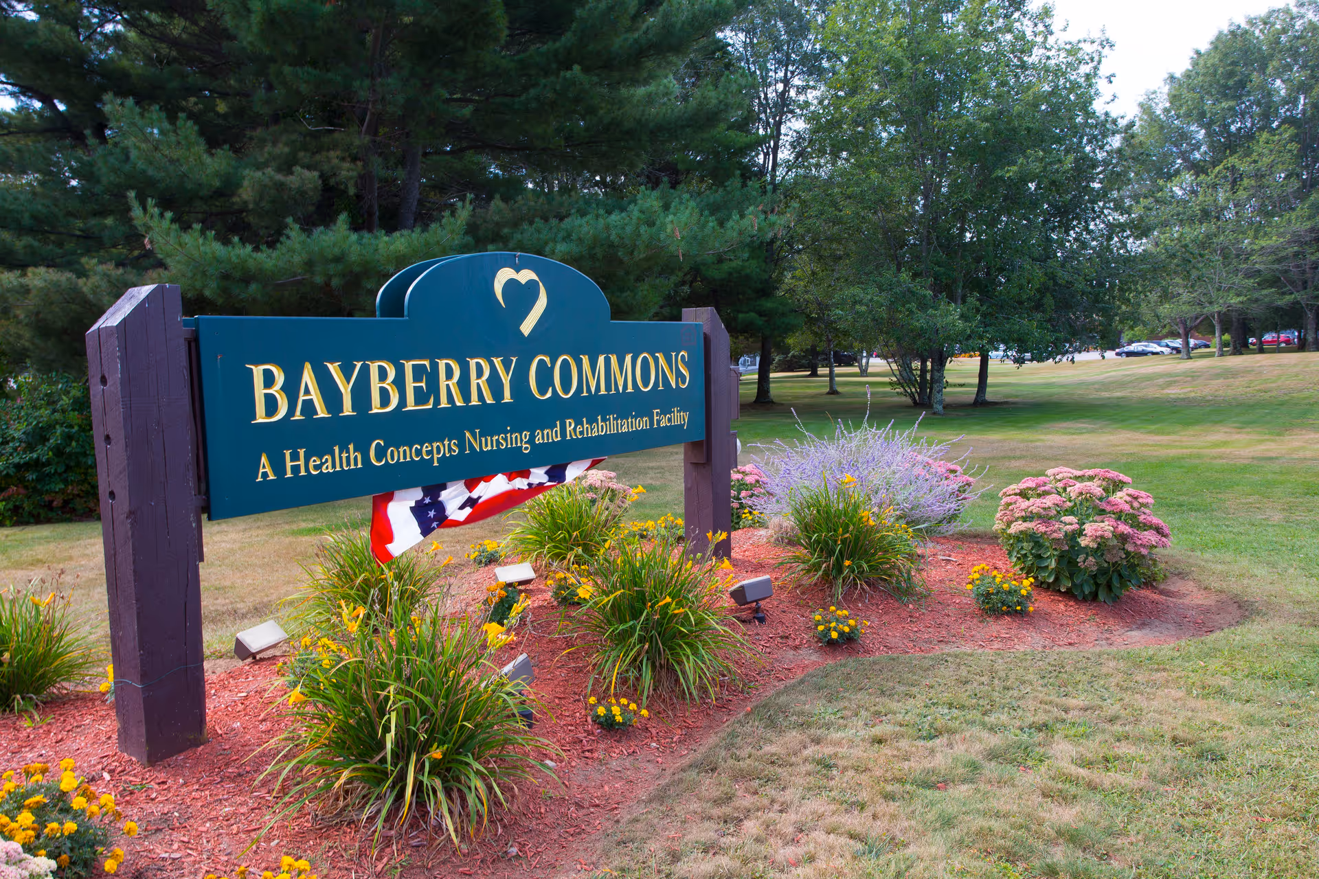 Outdoor view of a green sign for Bayberry Commons, a Health Concepts Nursing and Rehabilitation Facility, surrounded by landscaped flower beds with yellow and purple flowers, set against a backdrop of trees and a grassy area.