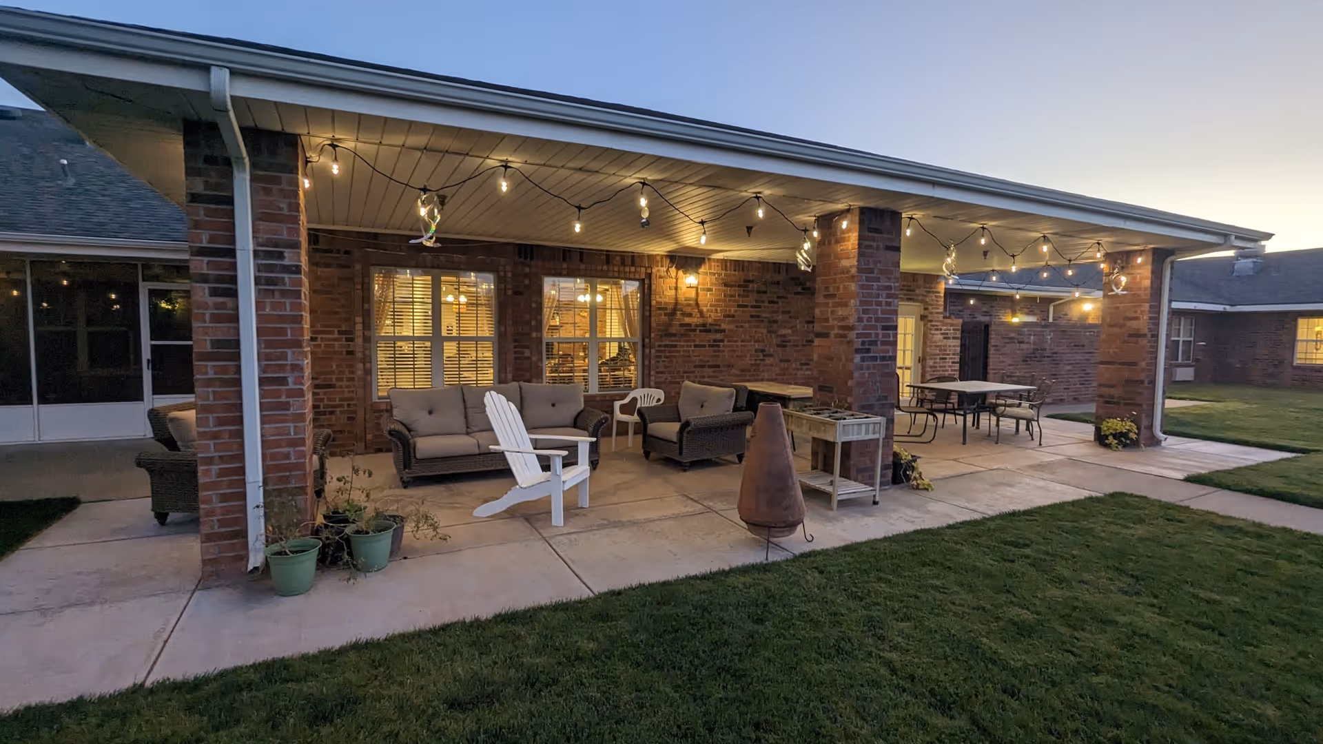 Covered outdoor patio with cushioned seating, tables, string lights, and a lawn in front of a brick building at dusk.