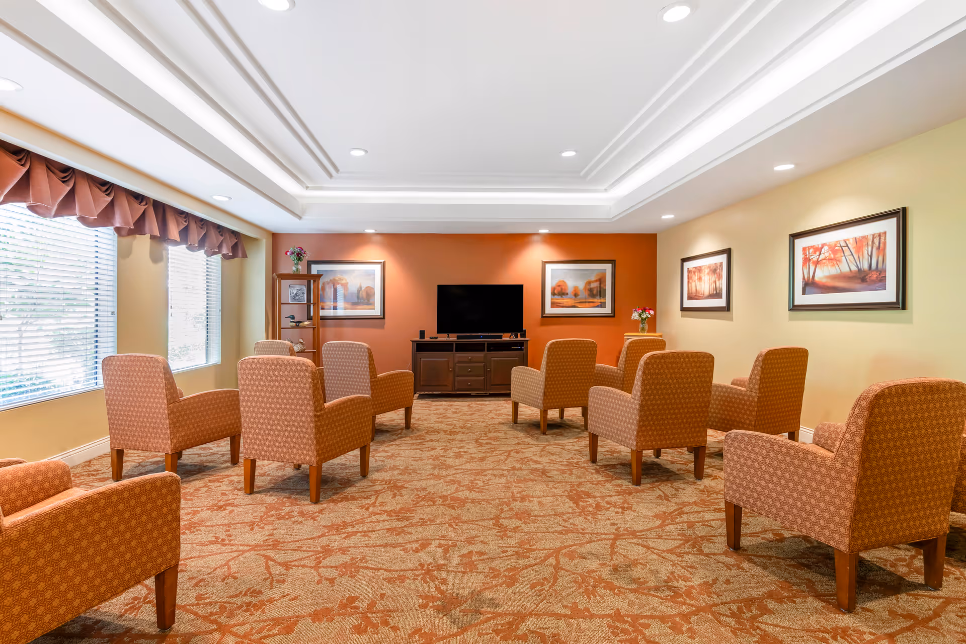 A bright communal living room with upholstered armchairs arranged facing a television and framed artwork on the walls.