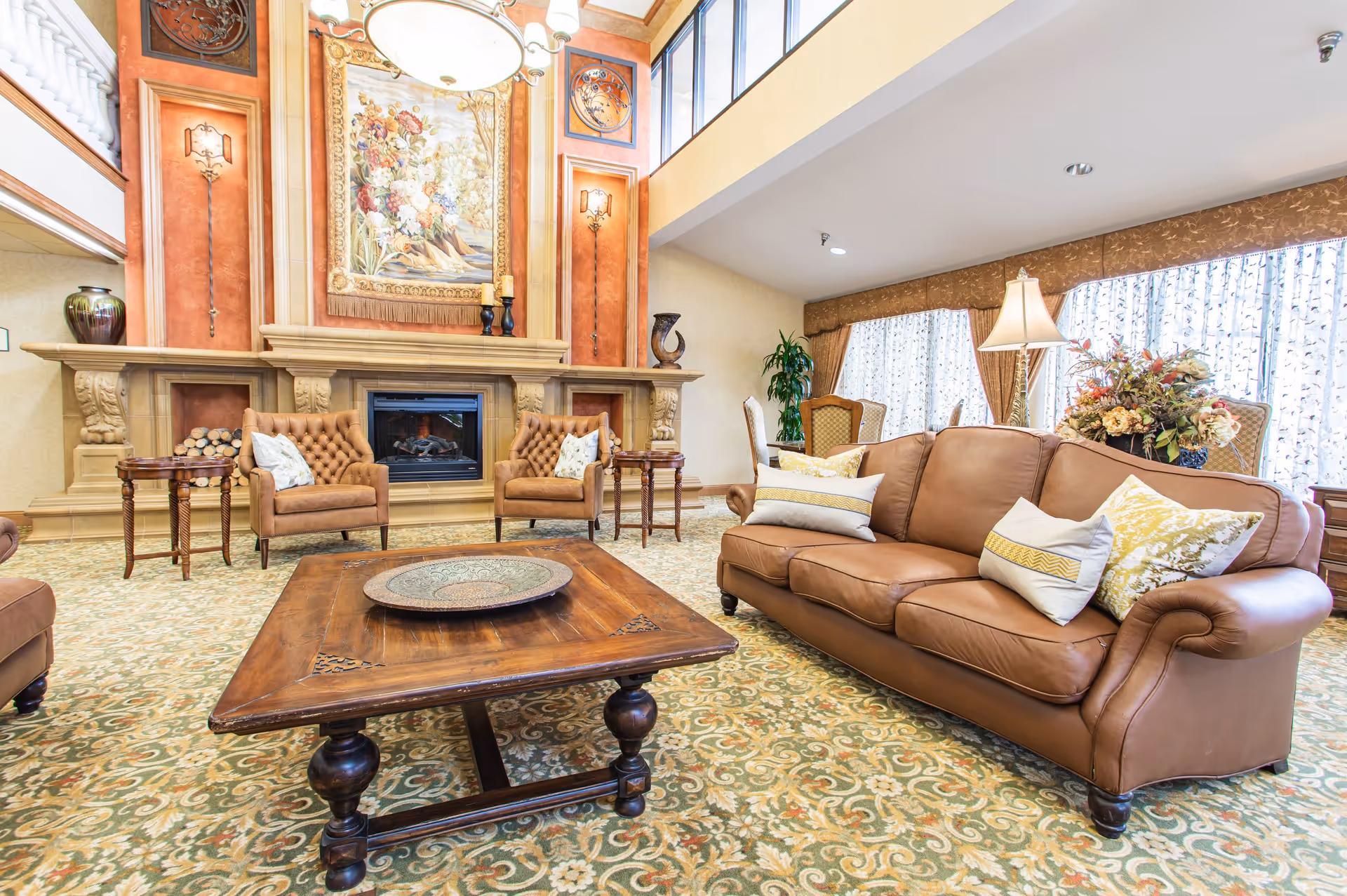 A spacious and elegant living room with a large brown leather sofa adorned with decorative pillows, two matching armchairs, a wooden coffee table with a decorative plate, and a fireplace with an ornate mantel. The room features a high ceiling, large windows with sheer curtains, patterned carpet, and warm-toned walls with artwork and sconces.