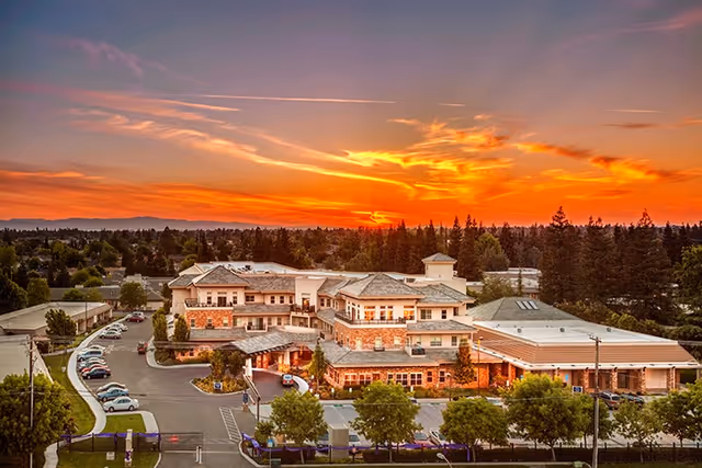 Aerial view of Covenant Living of Turlock senior living facility at sunset, showing a large multi-story building surrounded by trees, parking lots, and a scenic sky with orange and purple hues.