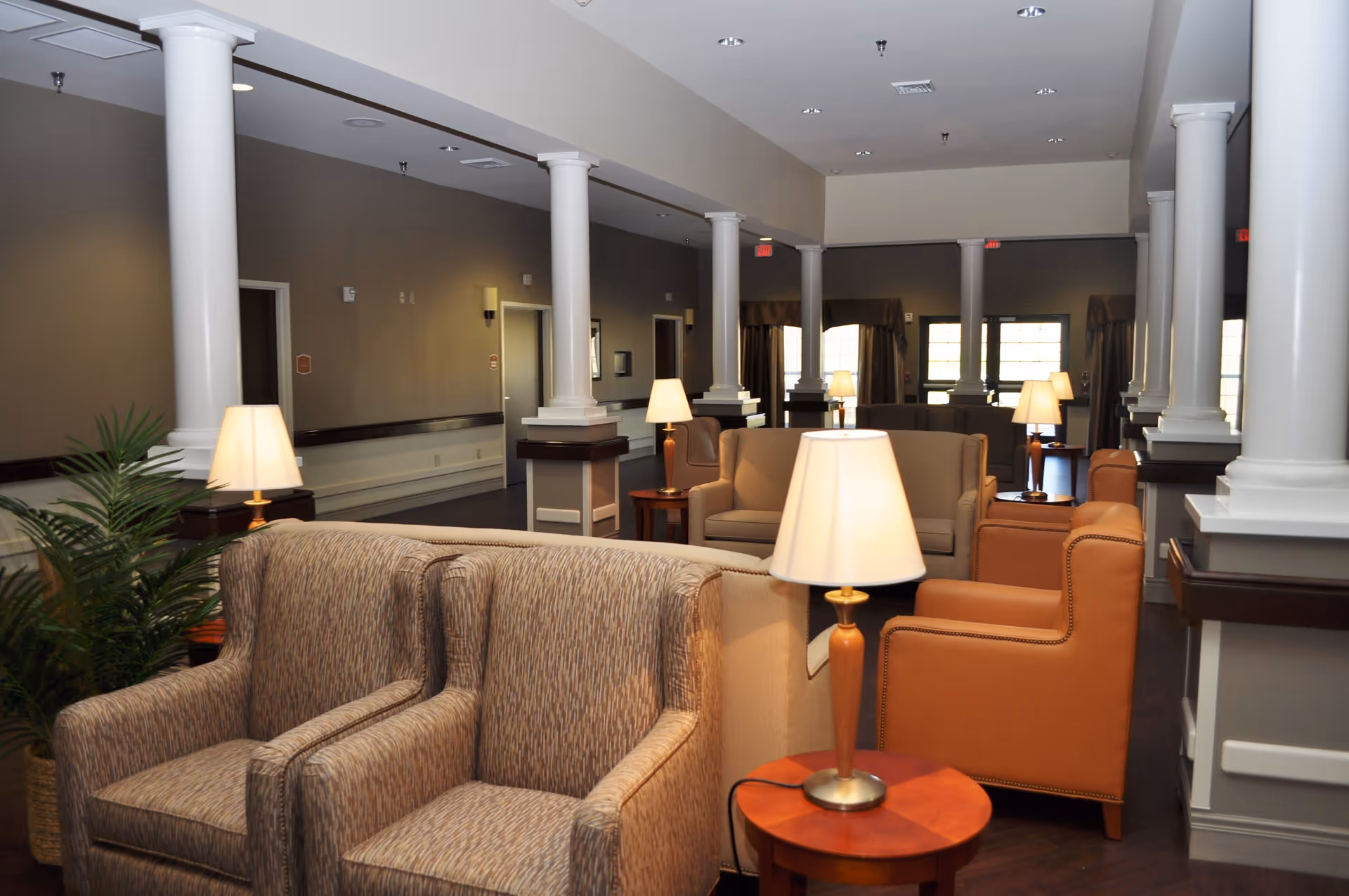 A spacious and well-lit common area with multiple armchairs and sofas arranged around small wooden tables with lamps. The room features white columns, dark wood flooring, and large windows with curtains in the background.