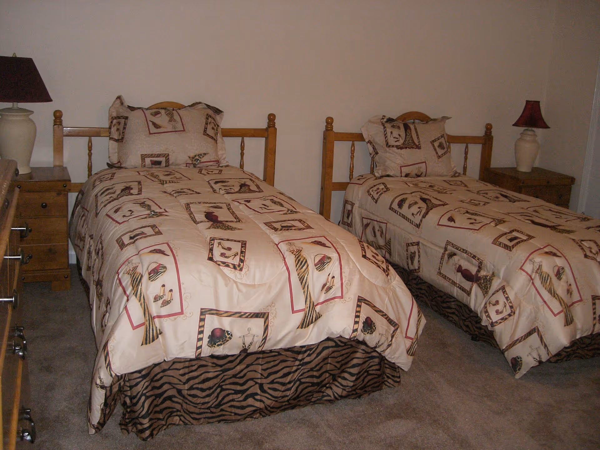 A bedroom with two twin beds featuring matching beige bedspreads with a pattern of hats and gloves. Each bed has a wooden headboard and a matching pillow. There are two wooden nightstands, each with a lamp, one with a white base and dark shade, the other with a white base and red shade. The room has beige carpet and plain light-colored walls.