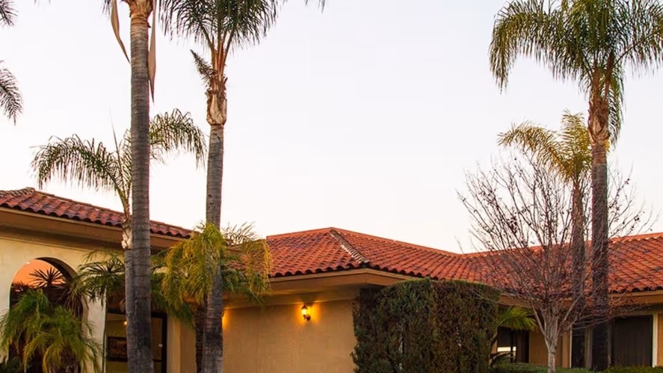 Front exterior of a Mediterranean-style building with a red tile roof, palm trees, shrubs, and wall sconces at dusk.