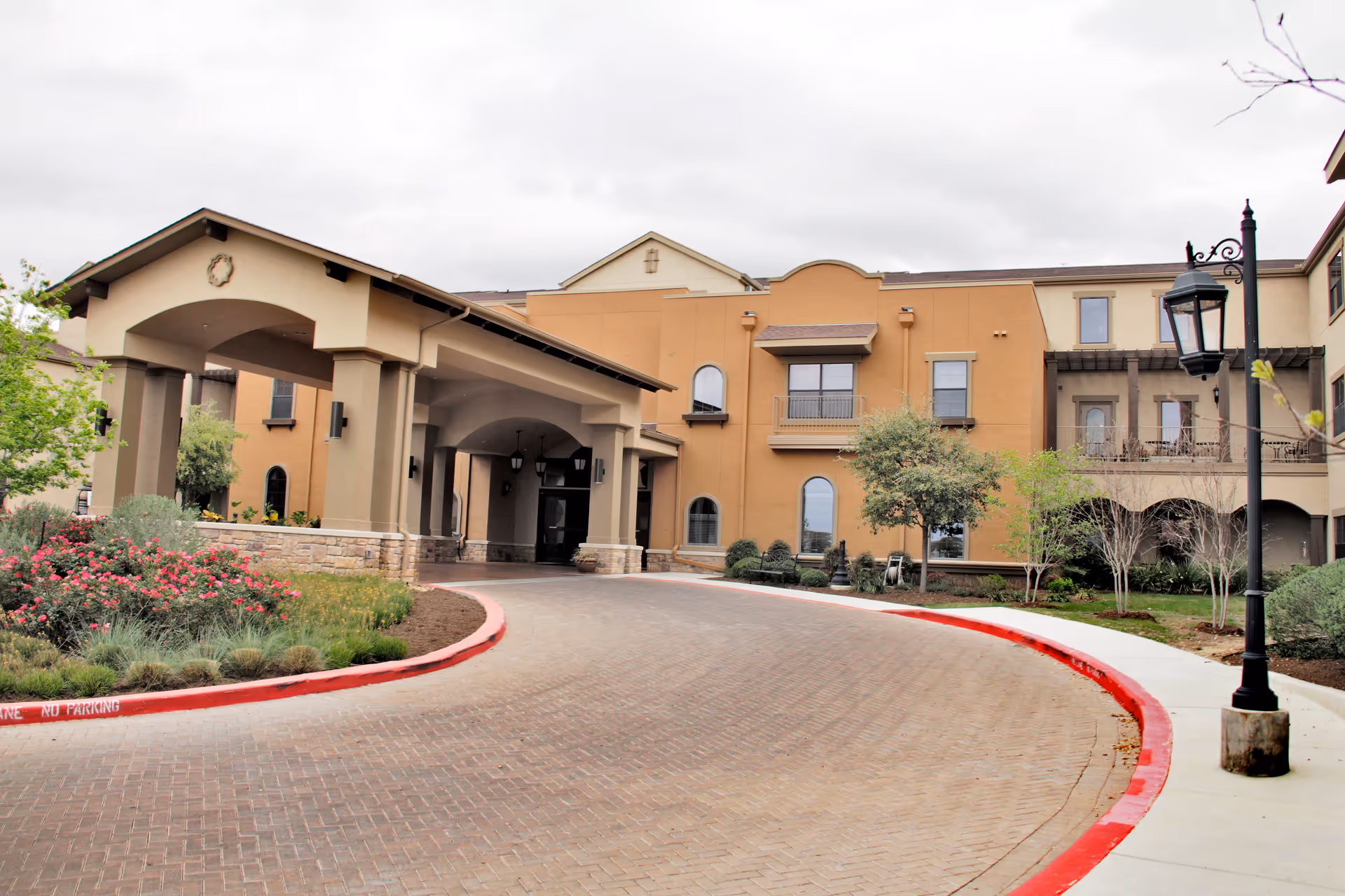Exterior view of Villa de San Antonio senior living facility showing the main entrance with a covered driveway, landscaped garden with flowers and shrubs, and a multi-story building with windows and balconies under a cloudy sky.