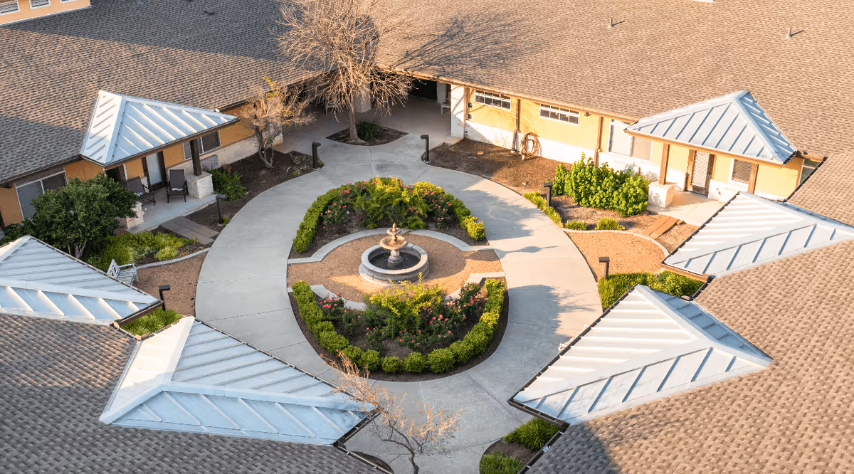 Aerial view of a courtyard in a senior living facility with a circular walkway surrounding a central garden featuring a small fountain and neatly trimmed bushes and flowers. The courtyard is enclosed by buildings with light brown walls and gray metal roofs, and there are some trees and outdoor seating areas visible.