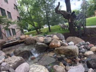 A landscaped outdoor area featuring a small rock waterfall and pond surrounded by various sized stones. In the background, there are trees, a grassy lawn, a metal bench, and part of a brick building visible on the left side.