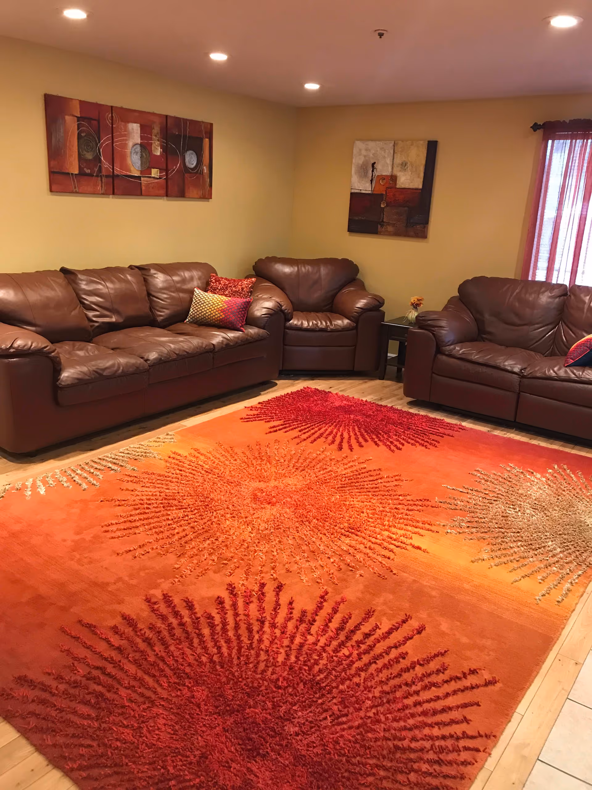 A cozy living room with three brown leather sofas arranged around a large orange area rug featuring red and beige sunburst patterns. The walls are painted yellow and decorated with two abstract paintings. A small side table with a flower vase is placed between two of the sofas. Recessed ceiling lights illuminate the room.