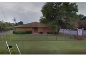 Single-story brick building with a brown roof surrounded by a black metal fence and green lawn. There is a large tree to the right of the building and a sign near the entrance. The sky is overcast.