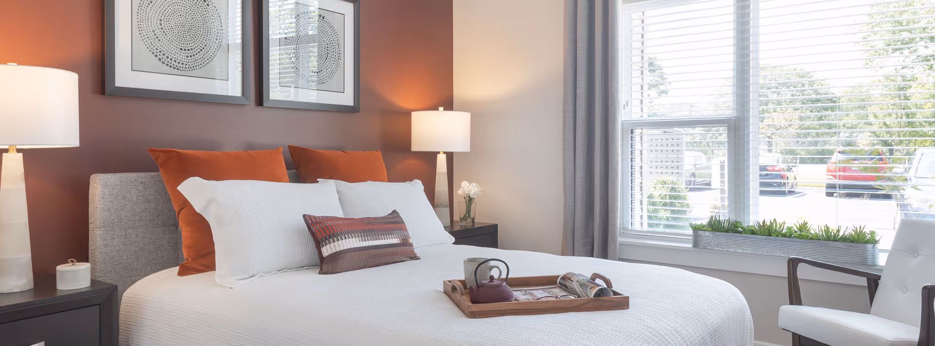 A neatly made bed with white bedding and orange accent pillows in a bedroom. There are two framed artworks on the wall above the bed, two bedside tables with lamps, and a window with blinds letting in natural light. A tray with a teapot and cup is placed on the bed.