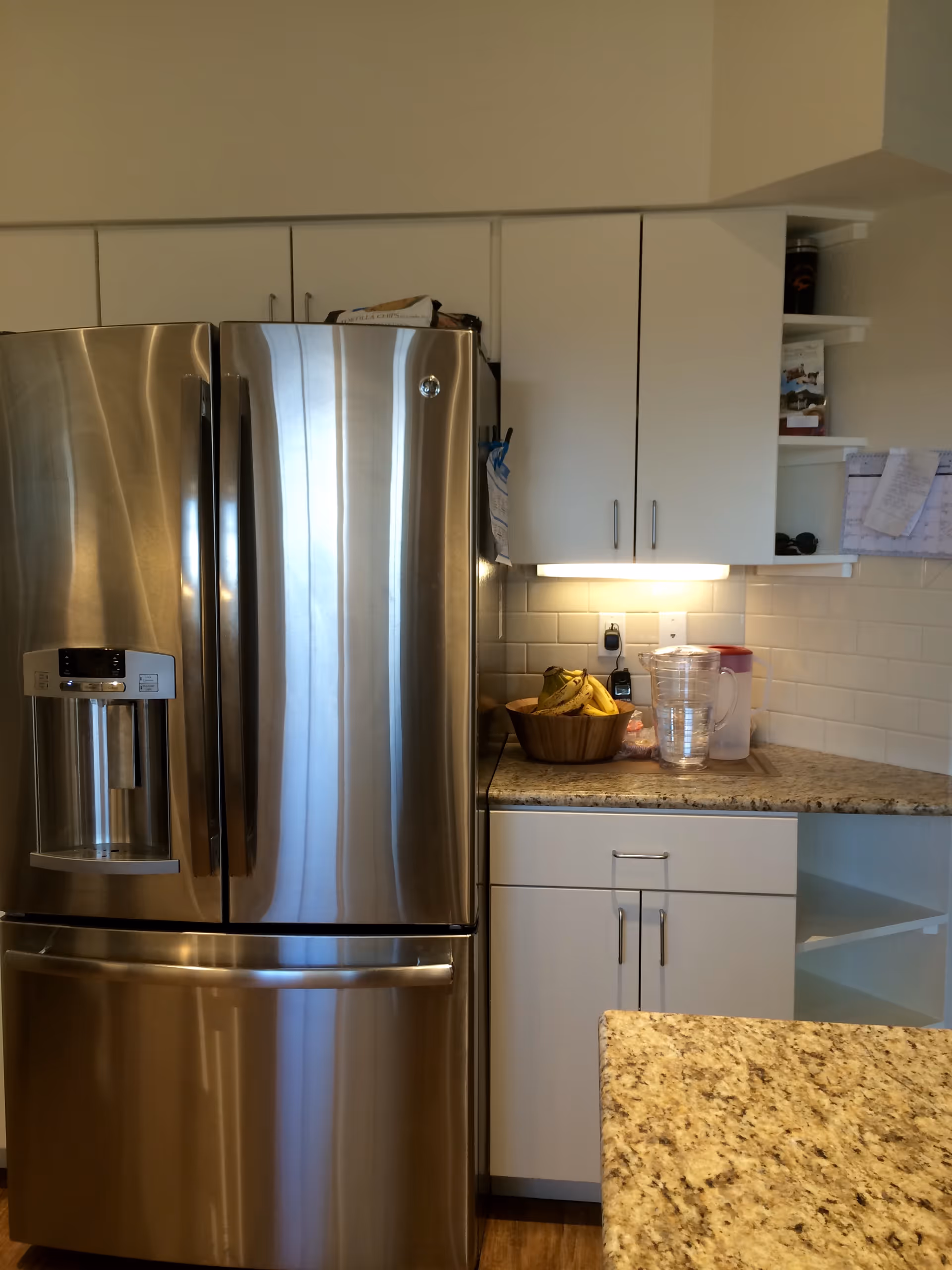 A modern kitchen corner featuring a stainless steel French door refrigerator with a water dispenser, white cabinets, granite countertops, a wooden bowl with bananas, a clear pitcher, and some papers pinned on the wall.