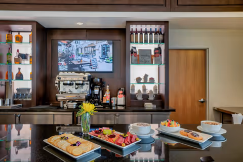A modern coffee and snack bar area with a coffee machine, cups, and various bottles on shelves. On the counter, there are plates with crackers, cheese, fruit, pastries, and two cups of coffee topped with foam. A small vase with a yellow flower is also on the counter. A flat-screen TV is mounted on the dark wood-paneled wall behind the counter, and a closed wooden door is visible to the right.