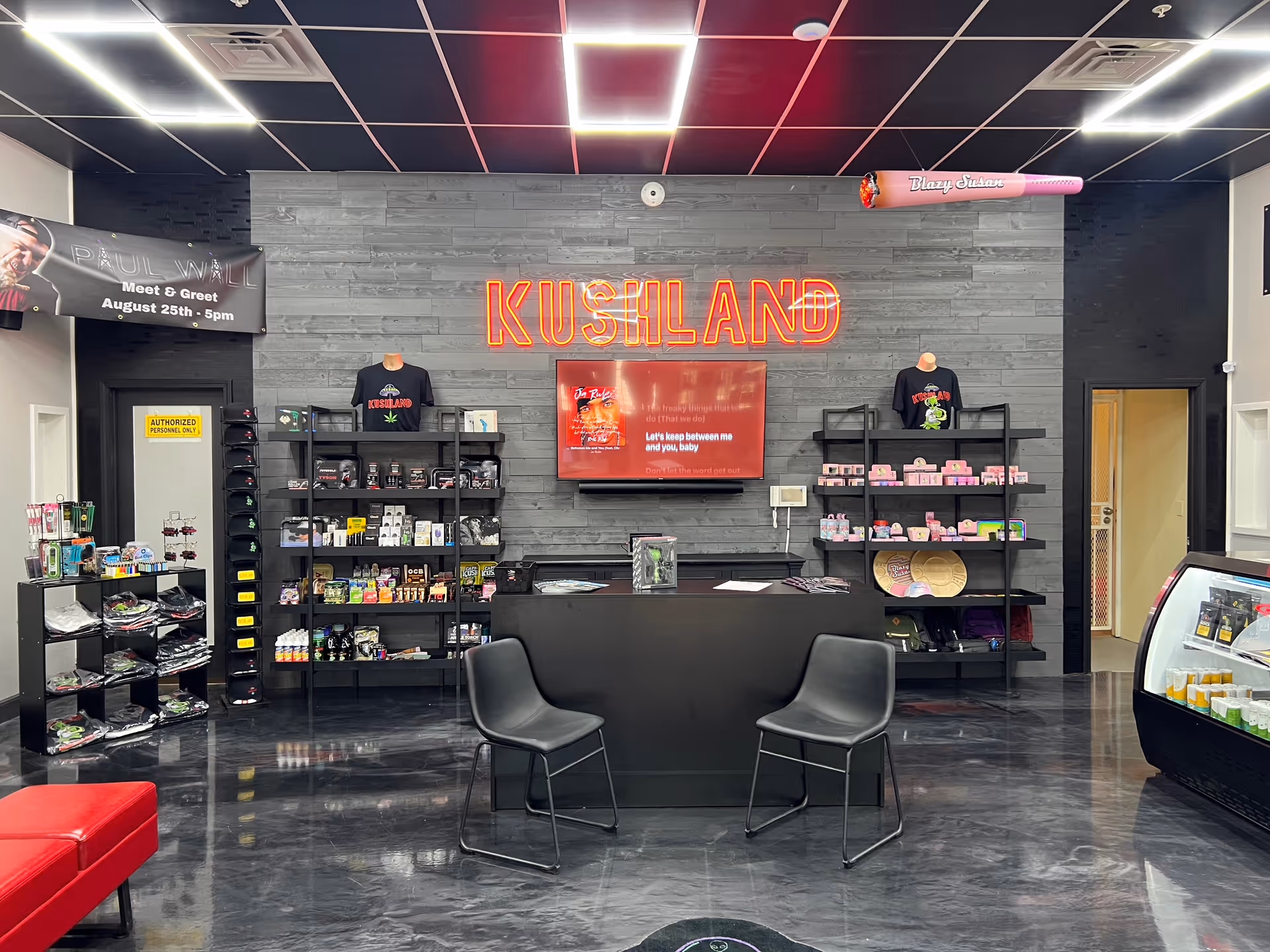 Interior view of a retail store with black and gray decor. The back wall features a neon sign that reads 'KUSHLAND' above a mounted TV screen. Shelves on either side display various products including apparel and packaged goods. Two black chairs are positioned in front of a black counter. The ceiling has a grid pattern with bright square lights. A banner on the left side announces a 'Paul Wall Meet & Greet August 25th - 5pm'.