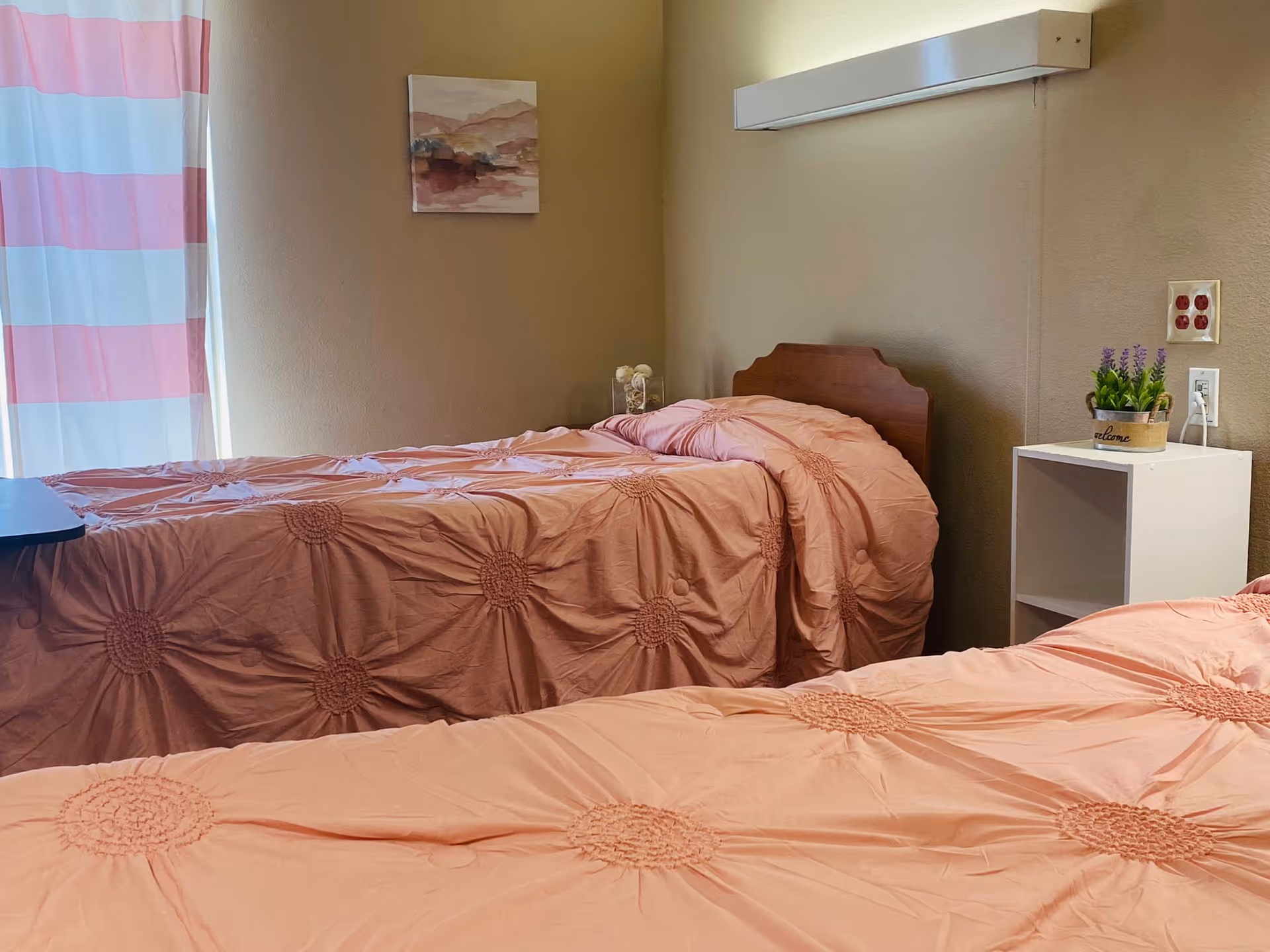 Two single beds with matching pink bedspreads in a small, simply furnished bedroom with a nightstand and window.