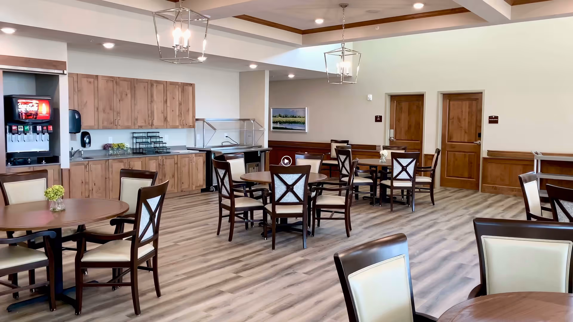 Bright dining room with round wooden tables and chairs, a serving counter with a beverage dispenser, and wood-paneled doors and trim.